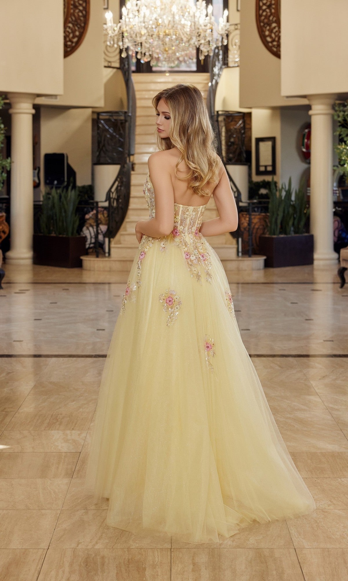 A woman wearing the Nox Anabel Q1773 long prom dress with 3D floral appliques stands indoors, facing away from the camera in a grand hall with a sweeping staircase and sparkling chandelier.